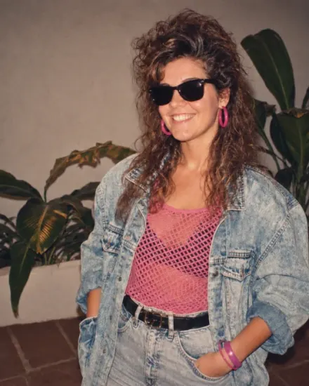 A smiling woman in black sunglasses, bright pink hoop earrings, a pink mesh top, and an oversized denim jacket poses in front of indoor plants. A red-orange date stamp reads “85 7 21” in the lower-right corner.