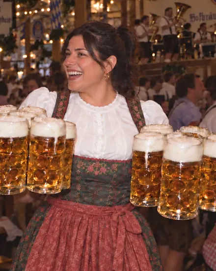 A smiling woman in a dirndl carries ten foaming beer steins through a crowded Oktoberfest tent.